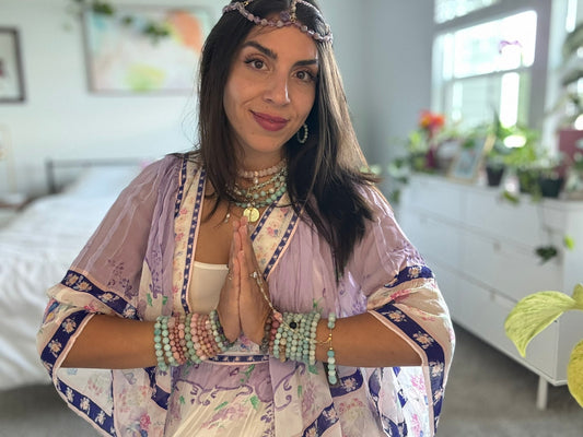 A mid-thirties brunette woman wearing way too much beaded gemstone jewelry, a flowery purple dress, with hands in a prayer pose.