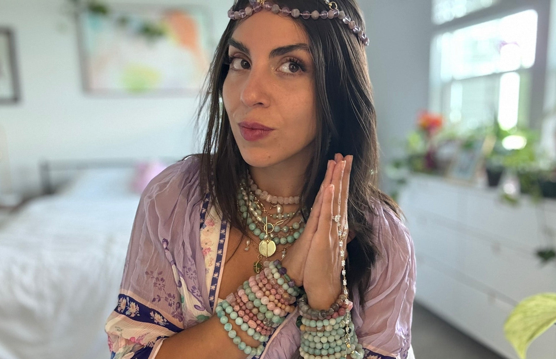 Woman in a room with a blurred background, wearing a patterned outfit and multiple necklaces.